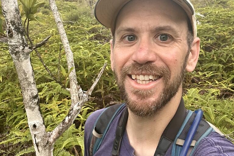 Smiling young man wearing basball cap, brown beard, Blue tshirt and jacket . Background forest of Madagascar with bushes and trees and a small bit of cloudy sky