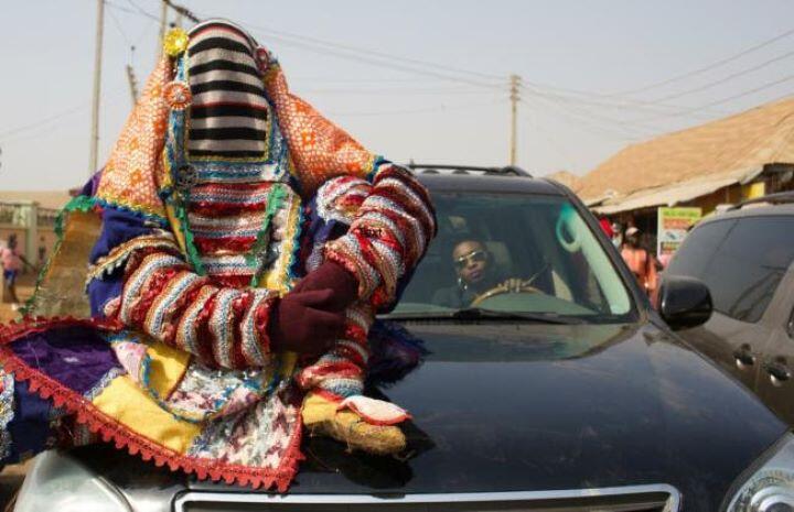 Still from film "Egúngún (Masquerade)". Man in head to toe colorful masquerade sitting on hood of car with driver with sunglasses sitting behind steering wheel on a street in a Nigerian town.